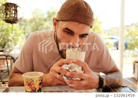 Man in a brown beanie eating a burrito wrapped in foil at a Mexican cafe table. Close front view portrait Man in a brown beanie eating a burrito wrapped in foil at a Mexican cafe table. Close front view portrait 132061459