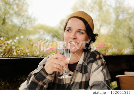 Woman smiling while holding and drinking a glass of water at an outdoor cafe table with flowers in background during autumn Woman smiling while holding and drinking a glass of water at an outdoor cafe table with flowers in background during autumn 132061515