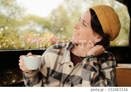 Smiling woman holding cup of coffee and looking aside while relaxing at outdoor cafe on an autumn day Smiling woman holding cup of coffee and looking aside while relaxing at outdoor cafe on an autumn day 132061516