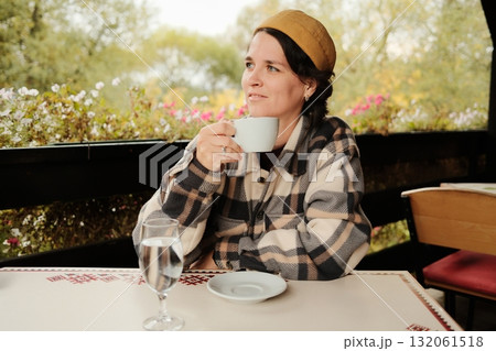 Woman in warm checkered jacket and hat sitting at outdoor cafe table drinking coffee and relaxing in cozy autumn atmosphere 132061518