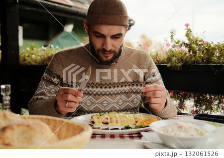 Man sitting at table with knife and fork looking at plate with traditional Balkan dish covered in cheese sauce, outdoor restaurant scene Man sitting at table with knife and fork looking at plate with traditional Balkan dish covered in cheese sauce, outdoor restaurant scene 132061526