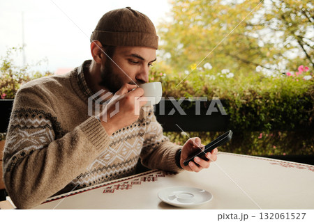 Young man in brown sweater holding coffee cup and using smartphone at outdoor cafe table, casual modern lifestyle scene Young man in brown sweater holding coffee cup and using smartphone at outdoor cafe table, casual modern lifestyle scene 132061527