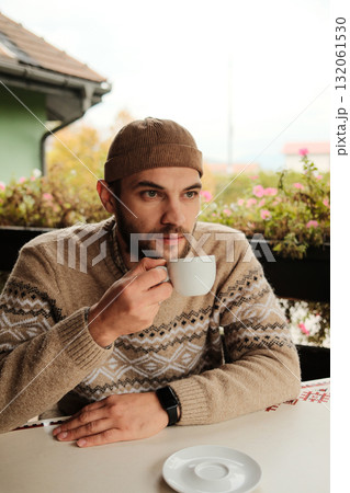 Man in warm sweater and hat drinking coffee at outdoor cafe table during autumn morning, calm and relaxed atmosphere Man in warm sweater and hat drinking coffee at outdoor cafe table during autumn morning, calm and relaxed atmosphere 132061530