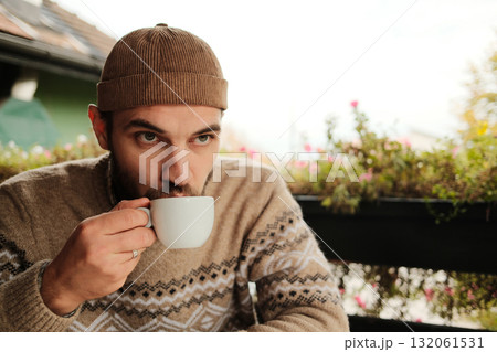 Man in warm sweater and hat drinking coffee at outdoor cafe table during autumn morning, calm and relaxed atmosphere Man in warm sweater and hat drinking coffee at outdoor cafe table during autumn morning, calm and relaxed atmosphere 132061531