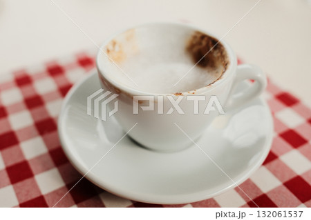 Close-up of a half-finished cappuccino with milk foam in a white cup on a red and white checkered tablecloth, cozy cafe detail 132061537