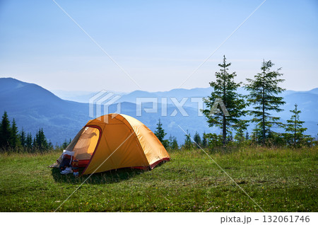 Bright yellow tent with camping gear on grassy hillside, overlooking breathtaking panorama of blue-tinged mountains. Young pine trees under clear blue sky, creating serene, picturesque camping spot. 132061746