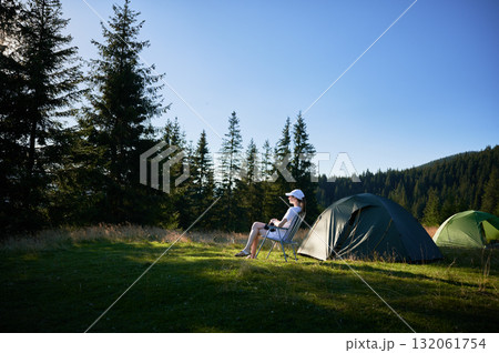 Woman camper relaxes next to tent with photo camera. Female tourist surrounded by tall evergreen trees and bathed in soft evening light, capturing peacefulness of moment. 132061754