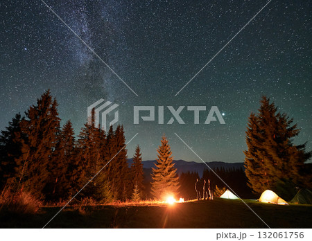 Night camping in mountains. Amidst forested mountain landscape, two tents glow warmly beside campfire under brilliant, star-filled sky. Silhouettes of tourists resting at campsite under Milky Way. 132061756