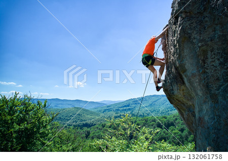 Aerial view of male rock climber ascending rugged limestone cliff with harness and rope for safety. Sportsman climbing on vertical large boulder at Dobvush Rocks in Carpathian mountains, Ukraine. 132061758
