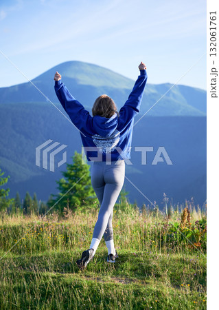 Woman tourist on grassy hill with beautiful mountain landscape in background. Young female wearing blue hoodie and grey leggings. Woman tourist on grassy hill with beautiful mountain landscape in background. Young female wearing blue hoodie and grey leggings. 132061761