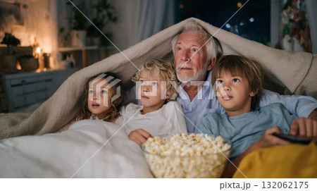 Grandfather and two grandchildren snuggled under a blanket, enjoying a movie night together, creating cherished family memories and emotional connections Grandfather and two grandchildren snuggled under a blanket, enjoying a movie night together, creating cherished family memories and emotional connections 132062175