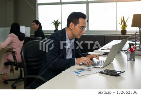 Focused businessman in formal suit working on laptop in modern office with colleagues in background Focused businessman in formal suit working on laptop in modern office with colleagues in background 132062536