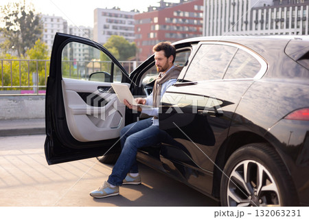 A man sits in a black car, working on a laptop with the door open, possibly in a parking lot. 132063231