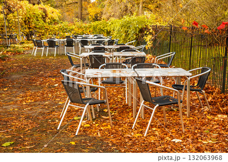 Empty outdoor cafe tables and chairs covered with autumn leaves in a park Empty outdoor cafe tables and chairs covered with autumn leaves in a park 132063968