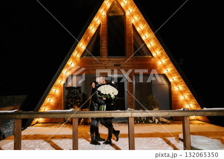 Romantic couple hugging and holding white roses bouquet in front of their chalet during winter night 132065350