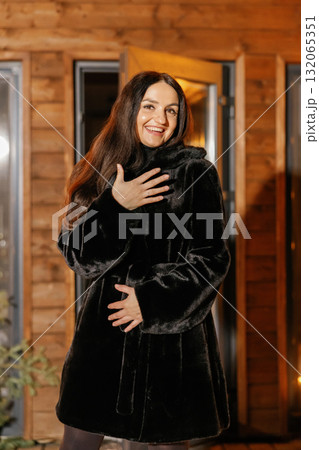 Happy woman wearing black fur coat and smiling near wooden house 132065351