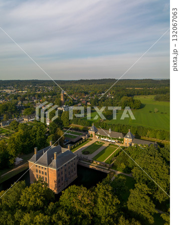 Aerial view of historic moated castle with gardens, bridge, and adjacent long building, surrounded by trees, water features, and a nearby village under warm sunlight. Aerial view of historic moated castle with gardens, bridge, and adjacent long building, surrounded by trees, water features, and a nearby village under warm sunlight. 132067063