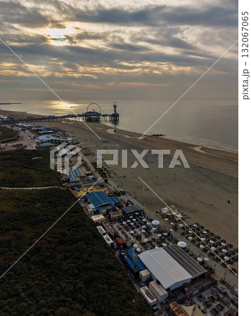 Aerial view of beachside pier Ferris wheel and tower rise over sea, umbrellas line sandy shore, with adjacent event tents, buildings, and sunlit reflections creating a vibrant coastal scene. 132067065