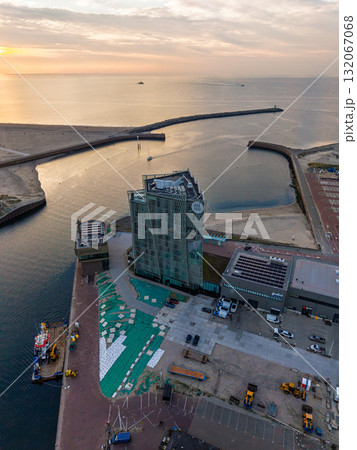 Aerial view of coastal harbor modern glass building near docks, construction equipment on site, breakwater protecting boats, sunset glow over sea blends urban design with maritime infrastructure. Aerial view of coastal harbor modern glass building near docks, construction equipment on site, breakwater protecting boats, sunset glow over sea blends urban design with maritime infrastructure. 132067068