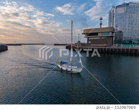 Aerial view of harbor white sailboat with blue cover glides through calm water, modern glass building on dock reflects warm sunset light, blending serene motion with sleek architecture. 132067069