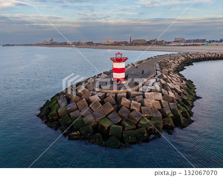 Aerial view of breakwater with red white lighthouse, people gathered near edge, concrete blocks protect harbor, coastal city and second tower in background under partly cloudy evening sky. 132067070
