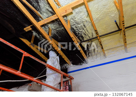 Contractor in protective gear applies refractory paint insulation using spray gun to wooden ceiling beams at construction site. 132067145
