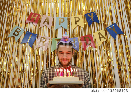 Man holding birthday cake with candles lit, golden backdrop and happy birthday banner behind 132068198
