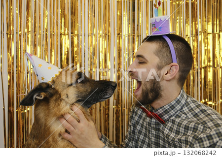 A playful man and his German Shepherd dog in party hats face each other during a fun birthday celebration. Pet birthday party concept 132068242