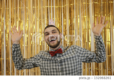 Smiling man in a party hat with hands raised, celebrating birthday against golden background. Birthday party concept 132068261