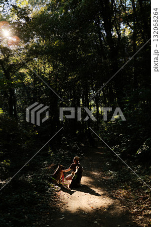 Woman kneeling on a forest path with two dogs German and Australian Shepherd, warm sunlight filtering through the trees, peaceful moment in nature. Travel and hiking with pets concept 132068264