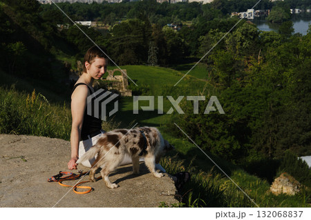Woman sits on cliff edge with red merle Border Collie puppy on leash, enjoying view of green valley and city 132068837