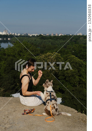 Woman trains red merle Border Collie puppy to give paws on cliff viewpoint overlooking river and Belgrade city skyline 132068838