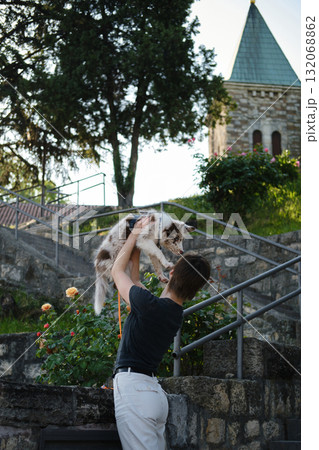 Woman lifts red merle Border Collie puppy in the air near flowers and stone wall, joyful playful outdoor moment. Travel with dog at old town fortress 132068862
