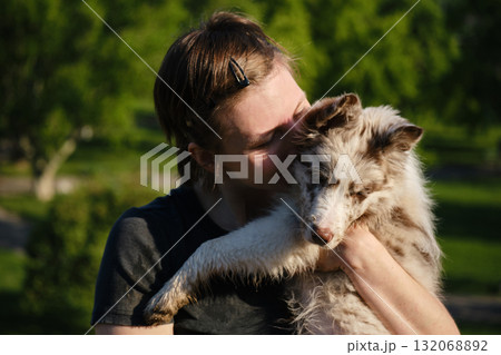 Smiling young woman holds red merle Border Collie puppy in arms outdoors, enjoying warm sunshine and green park background 132068892