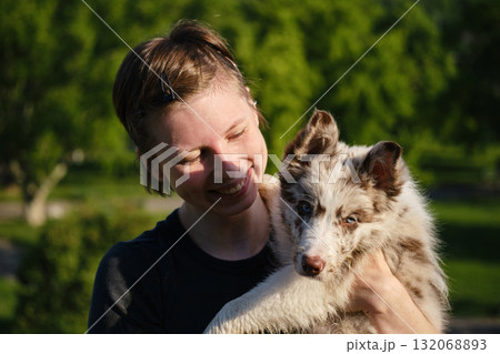 Smiling young woman holds red merle Border Collie puppy in arms outdoors, enjoying warm sunshine and green park background Smiling young woman holds red merle Border Collie puppy in arms outdoors, enjoying warm sunshine and green park background 132068893