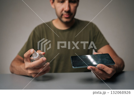 Selective focus portrait of man spraying sanitizer onto mobile phone screen, highlighting importance of hygiene and disinfection in everyday life on gray table surface. Concept of phone maintenance 132068926