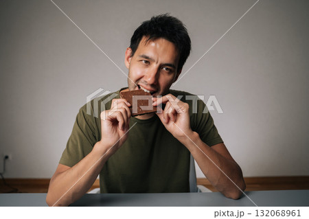 Portrait of happy Caucasian man sitting at table biting chocolate, savoring rich flavor of dark chocolate bar, blissful enjoyment as takes satisfying and delicious bite, on isolated background. Portrait of happy Caucasian man sitting at table biting chocolate, savoring rich flavor of dark chocolate bar, blissful enjoyment as takes satisfying and delicious bite, on isolated background. 132068961