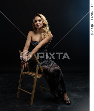 Blond fashion model posing on wooden chair in studio with dark background Blond fashion model posing on wooden chair in studio with dark background 132069217