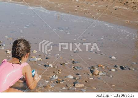 Little girl in a pink swimsuit playing actively with a red toy on the wet sand, building a moat and fort on a sunny pebble beach during a summer vacation Little girl in a pink swimsuit playing actively with a red toy on the wet sand, building a moat and fort on a sunny pebble beach during a summer vacation 132069518