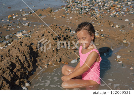 A happy little girl smiling and enjoying playing in the wet sand and shallow water on a pebble beach, sitting next to her handmade sand fort on a sunny day 132069521