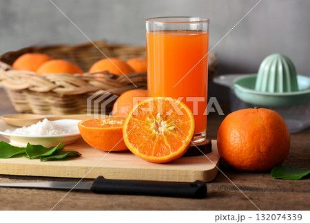 Half an orange is placed on a wooden chopping board. Placed on an old wooden table prepared for making orange juice, In the back there are oranges in the basket and the juicer container. Half an orange is placed on a wooden chopping board. Placed on an old wooden table prepared for making orange juice, In the back there are oranges in the basket and the juicer container. 132074339