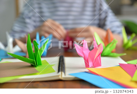 A close up shot of a bird folded from colorful origami paper with a blurred background of a woman folding the paper on wooden table top A close up shot of a bird folded from colorful origami paper with a blurred background of a woman folding the paper on wooden table top 132074343
