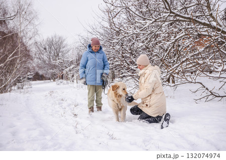 Elderly woman and her adult daughter enjoying a snowy day outdoors with their Golden Retriever. The daughter kneels to pet the dog while the mother stands nearby holding the leash amid snow-covered Elderly woman and her adult daughter enjoying a snowy day outdoors with their Golden Retriever. The daughter kneels to pet the dog while the mother stands nearby holding the leash amid snow-covered 132074974