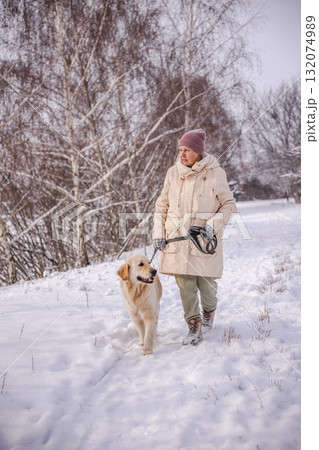Elderly woman walking her Golden Retriever through a snowy countryside path. The woman smiles warmly at the dog while holding the leash, surrounded by snow-covered trees on a peaceful winter day. 132074989