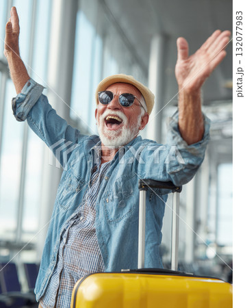 Ecstatic senior man cheering with arms up in a bright airport terminal next to a yellow suitcase. Represents joyful travel, retirement, adventure, happy holidays, and active senior lifestyle 132077983