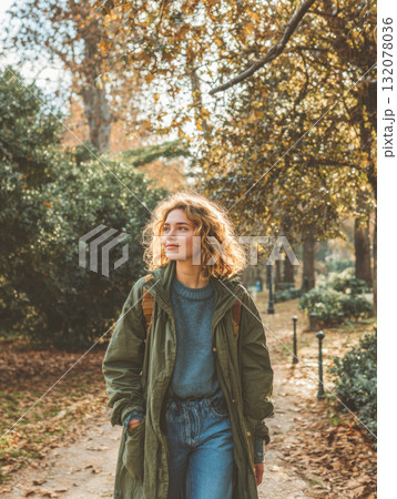 A peaceful young woman with curly, sunlit hair is seen from the waist up, happily walking through a leafy autumn park, wearing a khaki trench coat and denim jeans, embodying nature, freedom 132078036