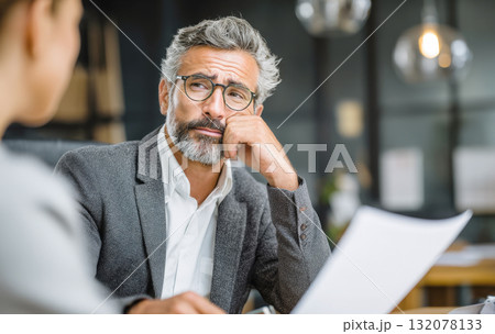 A mature businessman with a grey beard and eyeglasses leans forward, resting his chin on his hand, displaying a skeptical or thoughtful expression while listening to colleague and reviewing paperwork 132078133