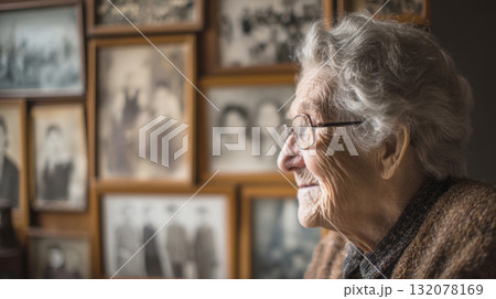 A senior woman with white hair and glasses smiles in profile, warmly gazing at a wall display of framed, vintage family photographs, capturing the nostalgia and joy of lifelong memories A senior woman with white hair and glasses smiles in profile, warmly gazing at a wall display of framed, vintage family photographs, capturing the nostalgia and joy of lifelong memories 132078169