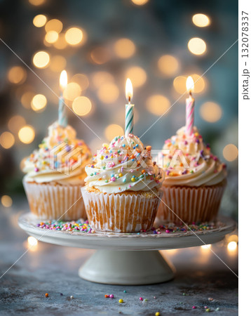 A close-up of three festive vanilla cupcakes topped with creamy white frosting, rainbow sprinkles, and lit birthday candles, presented on a white pedestal stand. A close-up of three festive vanilla cupcakes topped with creamy white frosting, rainbow sprinkles, and lit birthday candles, presented on a white pedestal stand. 132078337