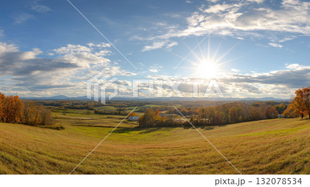 Stunning panoramic view of a rural valley in autumn, featuring golden fields, rolling hills, and forests ablaze with fall color under a brilliant sunburst and a wide, partly cloudy blue sky 132078534